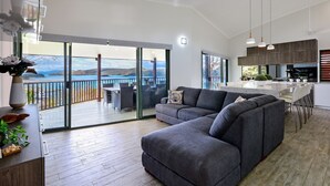 A modern living room with a grey sectional sofa, a dining area with a table and chairs, a kitchen with dark wood cabinets, and a balcony with a view of the sea.