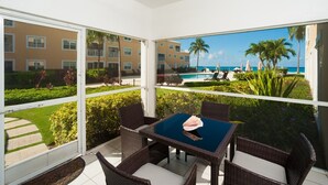A sunlit room with a glass table and wicker chairs, overlooking a pool and a building with balconies. - Regal Beach Club #612 (Seven Mile Beach)