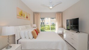 A hotel room with a large bed, a TV, a desk, and a balcony with a view of greenery. - Regal Beach Club #612 (Seven Mile Beach)