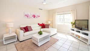 A modern living room with a white sofa, a glass coffee table, a TV on a white stand, and artwork on the wall. - Regal Beach Club #612 (Seven Mile Beach)