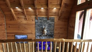 Un interior de madera con chimenea de piedra, vigas de madera y una pantalla de televisión mostrando un paisaje urbano.
