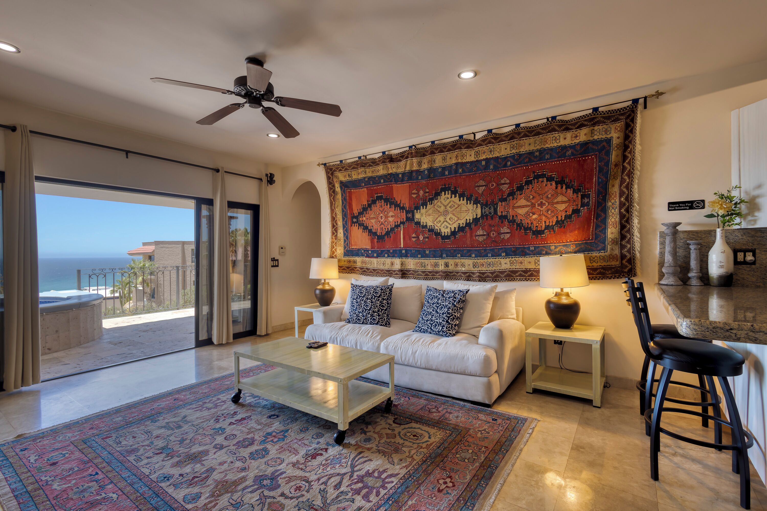 A spacious living room with a large window overlooking the ocean, a ceiling fan, a patterned rug, and a white sofa with blue pillows.
