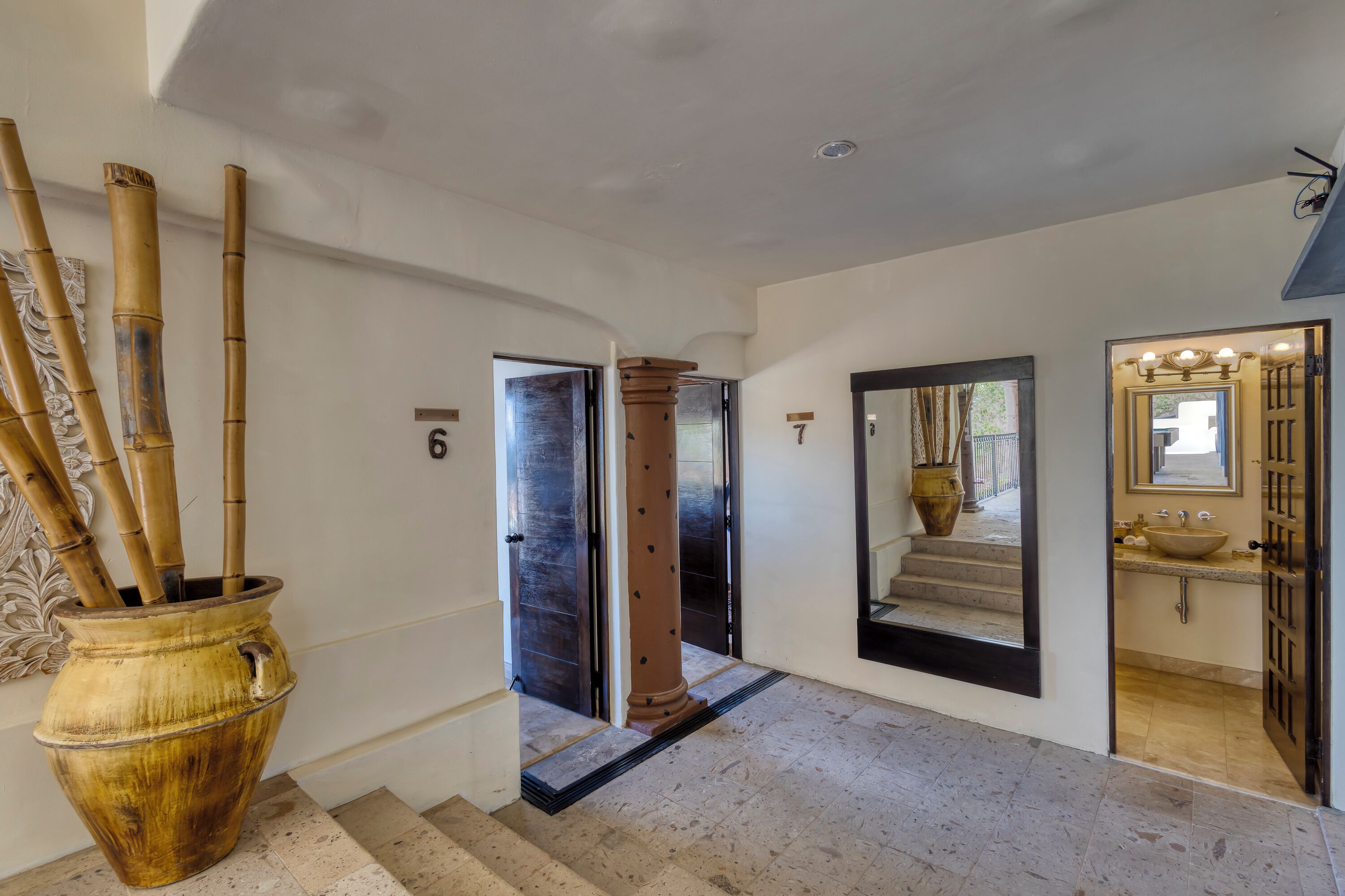 Un couloir avec une grande plante en bambou, un miroir et une salle de bain équipée d’un lavabo et d’un miroir.