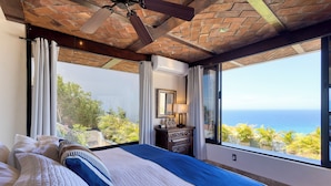 A bedroom with a large bed, a ceiling fan, a dresser, and a view of the ocean.