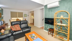 Un salon avec un canapé en cuir noir, une petite table basse avec des magazines, une étagère en osier ornée d’objets décoratifs et un téléviseur posé sur un meuble.
