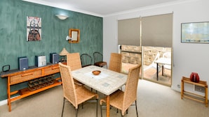 Un espace repas comprenant une table et des chaises, une console avec des haut-parleurs et une lampe, et une vue sur une terrasse extérieure accessible par une porte-fenêtre coulissante.