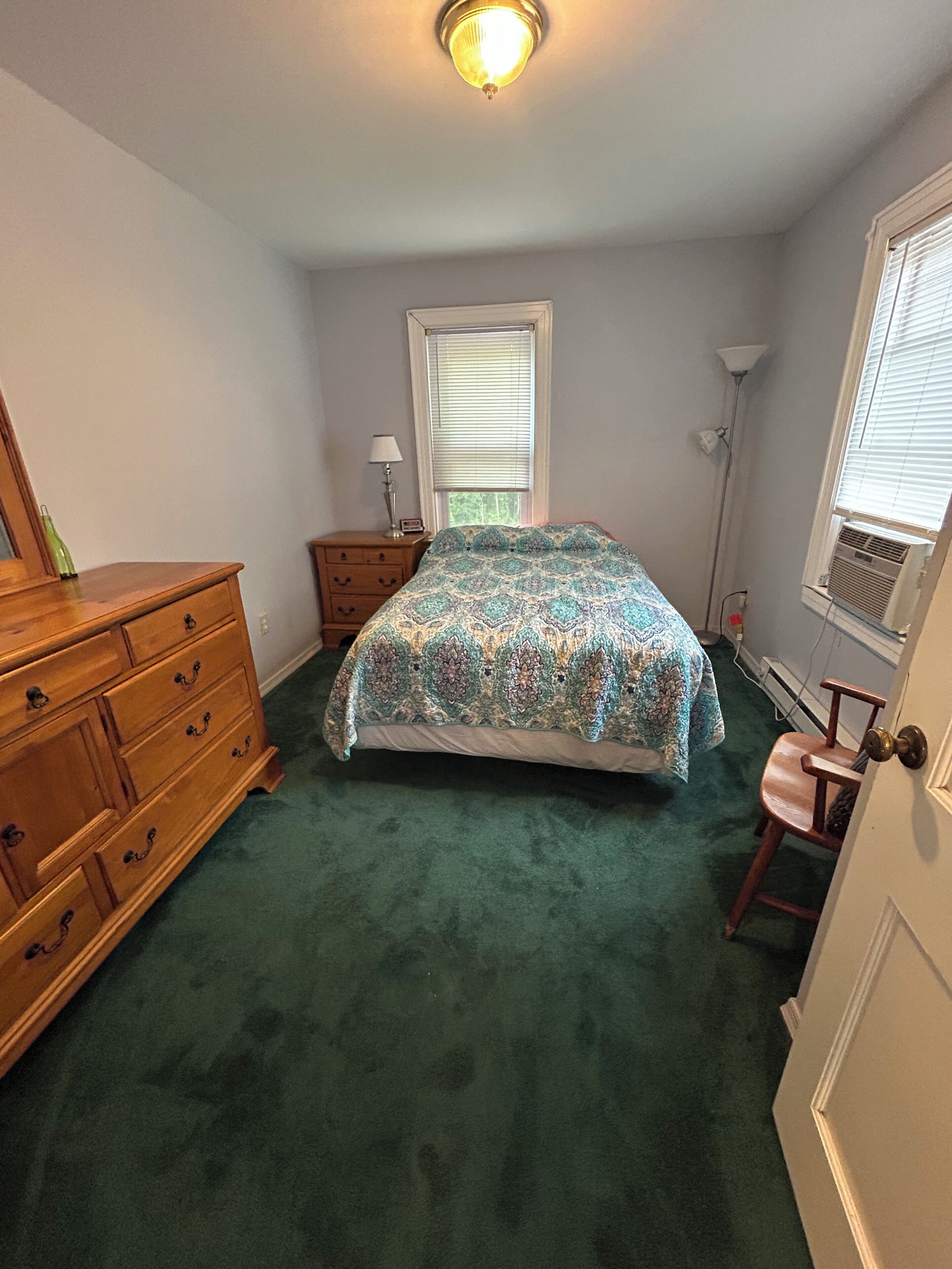 Une chambre à coucher comprenant un lit, une commode, une table de chevet, une lampe et une fenêtre avec des stores.
