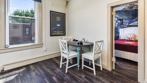 Une pièce comprenant une table à manger et des chaises, une fenêtre donnant sur un bâtiment en briques, et une porte menant à une chambre avec un lit et un mur décoré sur le thème de la galaxie.