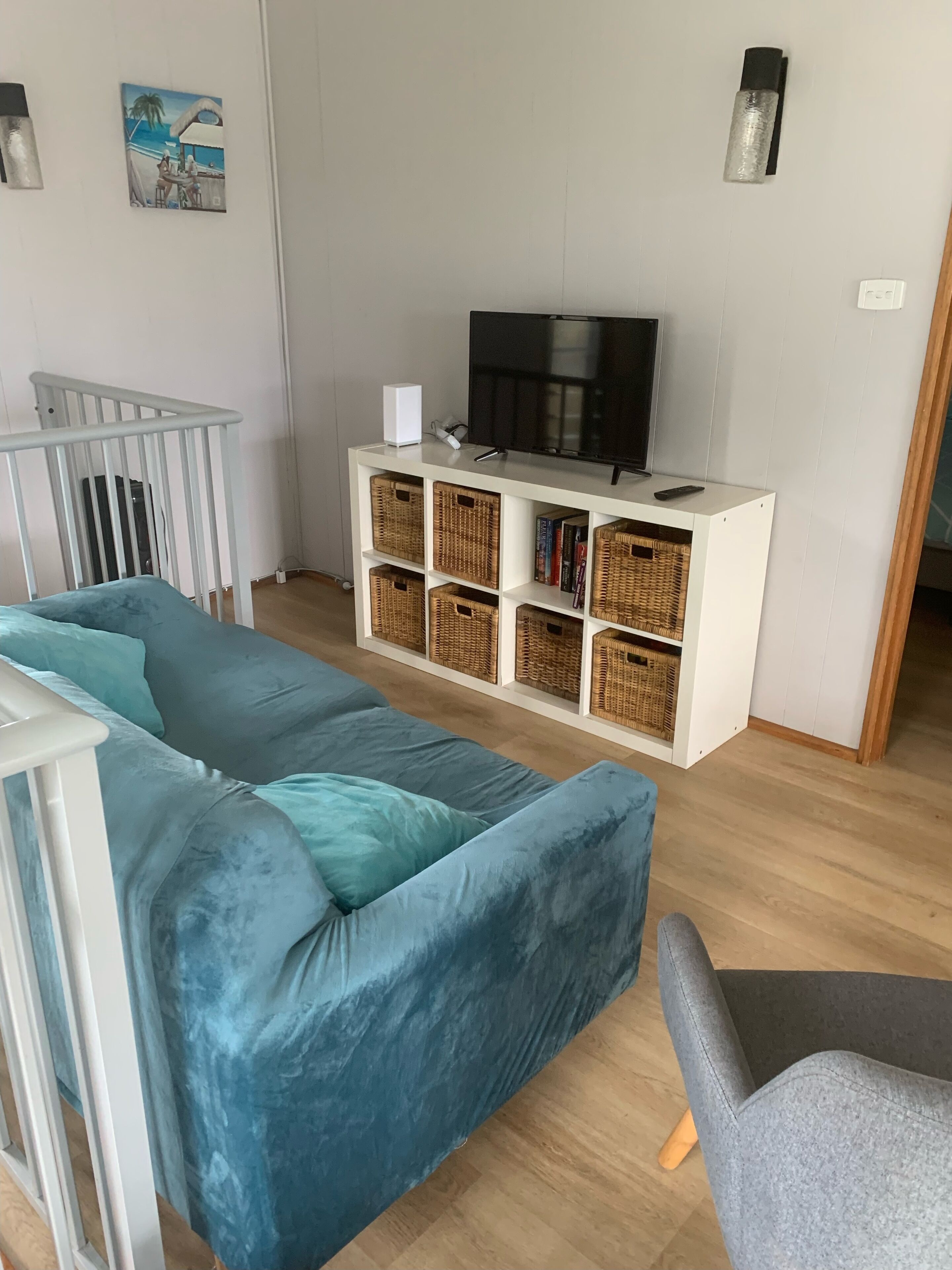 Een woonkamer met een blauwe bank, een witte meubelset met rieten manden, een televisie en een kleine tafel.