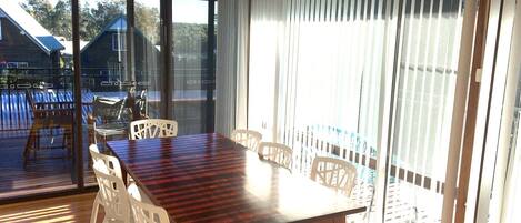 Un espace repas avec une table en bois et des chaises blanches, de grandes fenêtres équipées de stores et une vue sur un immeuble et des arbres à l’extérieur.