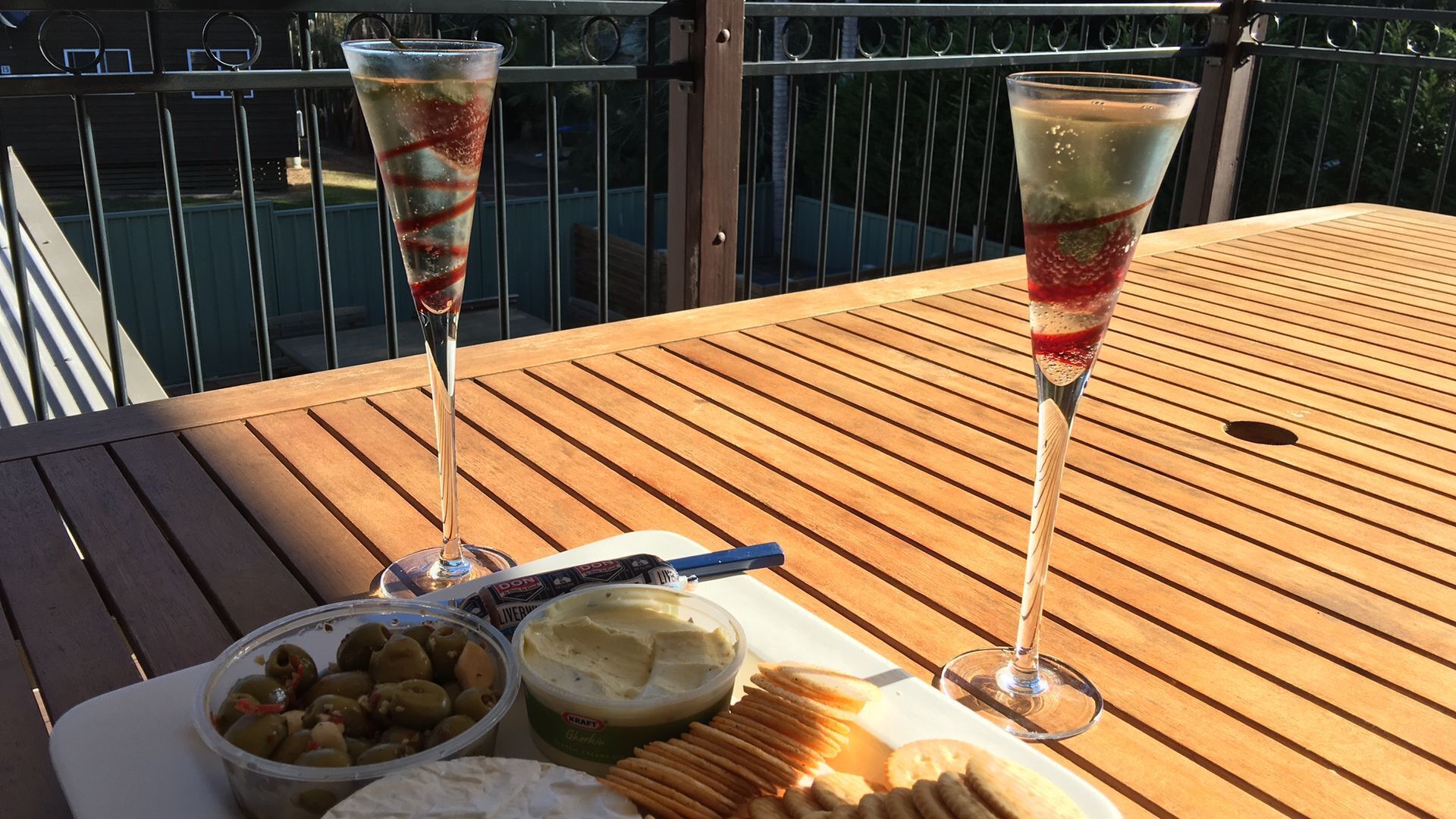 Two champagne glasses on a wooden table with olives, cheese, crackers, and a bottle of beer.