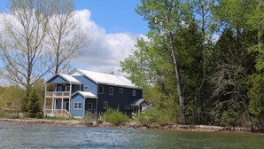 A two-story house with a blue exterior and a white roof, located near a body of water with trees in the background. - Loving Life at the Lake (Black River)