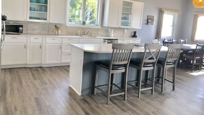 A modern kitchen with white cabinetry, a central island with bar stools, and a dining area with a large table and chairs. - Loving Life at the Lake (Black River)