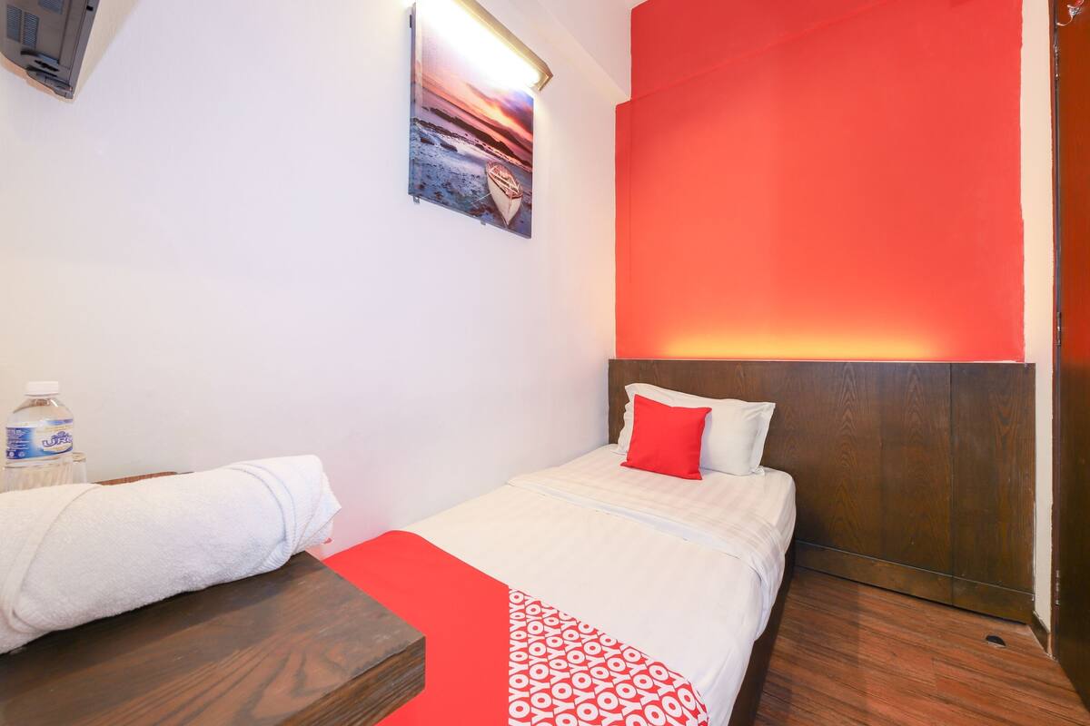Een hotelkamer met een bed, een houten nachtkastje, een rode en witte gebogen plaid, een witte handdoek, een fles water en een ingelijst foto aan de muur.
