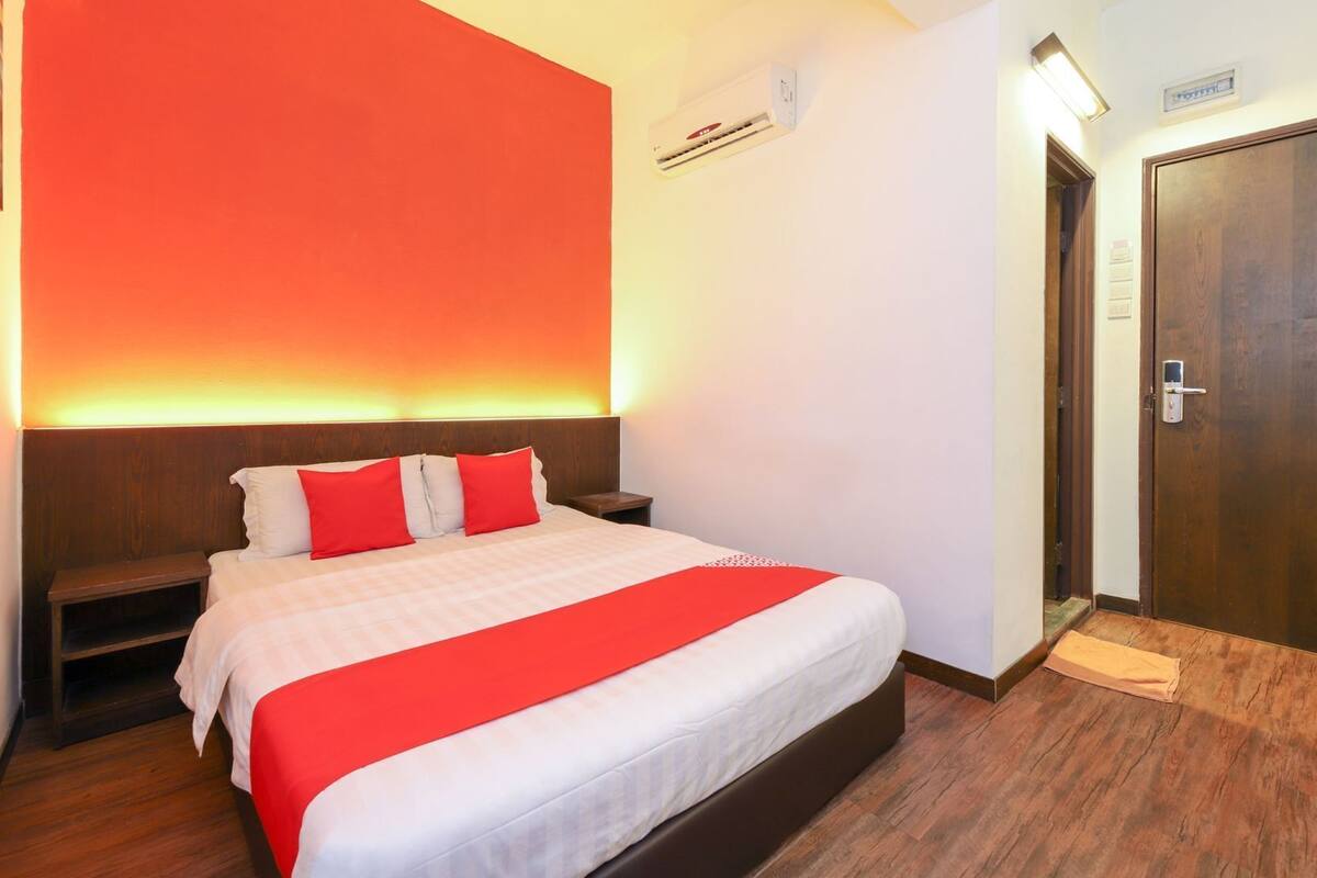 Een hotelkamer met een bed, nachtkastje, rode accentmuur, houten deur en een wandairconditioner.