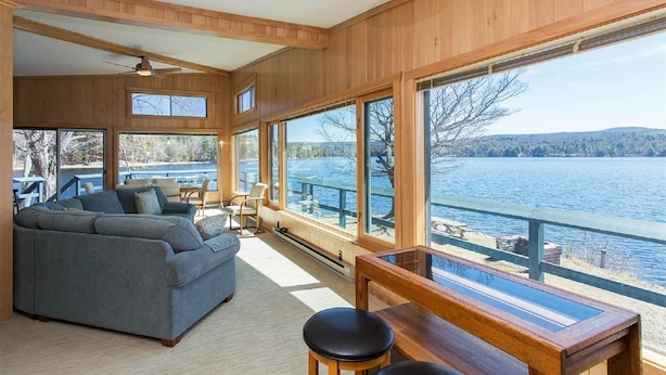 Uma sala de estar ampla com uma grande janela com vista para um lago, um ventilador de teto e um deck de madeira no exterior.