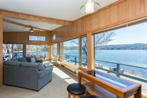 A spacious living room with a large window overlooking a lake, a ceiling fan, and a wooden deck outside. - Direct Waterfront! 230ft of frontage. 6BRs, 2BAs and beautiful views! (Hadley)