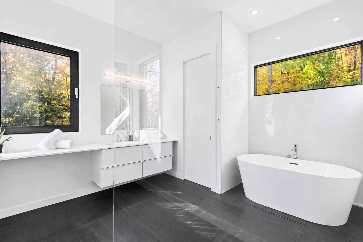 A modern bathroom with a freestanding bathtub, a large mirror, and a window with a view of trees.