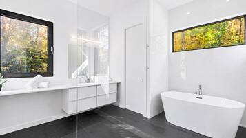 A modern bathroom with a freestanding bathtub, a large mirror, and a window with a view of trees.