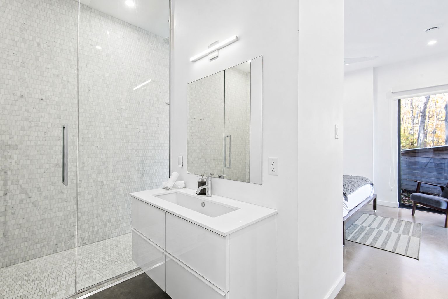 Une salle de bain moderne avec une douche à l’italienne en verre, un meuble-lavabo blanc doté d’un grand miroir, et une vue sur une chambre avec un lit et un fauteuil.