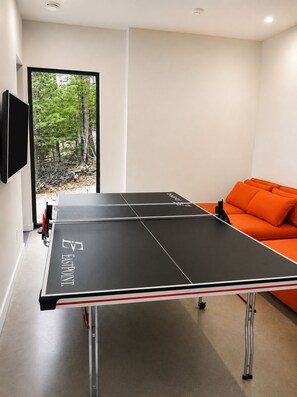 A ping pong table with a branded logo, a sofa with orange cushions, and a television mounted on the wall. - Architect-Designed Lakefront Villa Hot Tub Sleeps 14 (Mansonville)