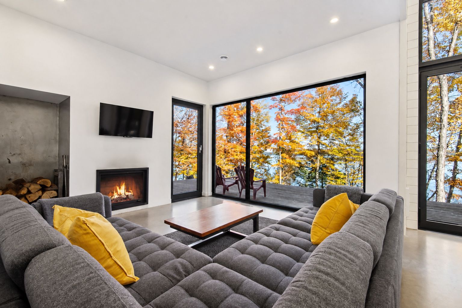 Un salon moderne avec une cheminée, des canapés gris, des coussins jaunes, une table basse en bois et de grandes fenêtres offrant une vue sur le feuillage automnal.