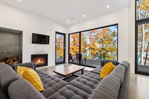 Een moderne woonkamer met een open haard, grijze banken, gele kussens, een houten salontafel en grote ramen met uitzicht op herfstbladeren.