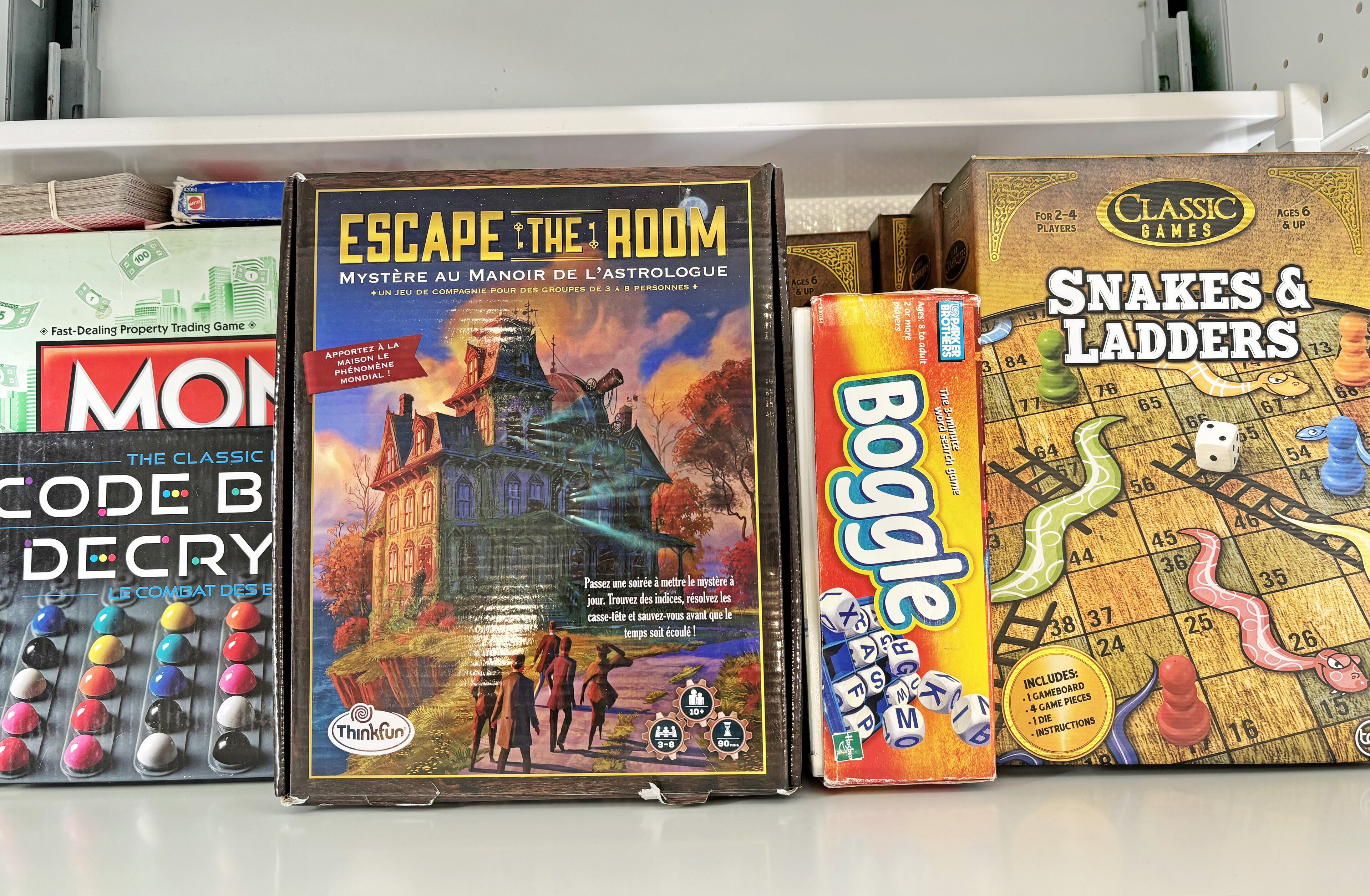 Uma estante com vários jogos de tabuleiro, incluindo 'Monopoly', 'Code B Decry', 'Escape the Room', 'Boggle' e 'Snakes & Ladders'.