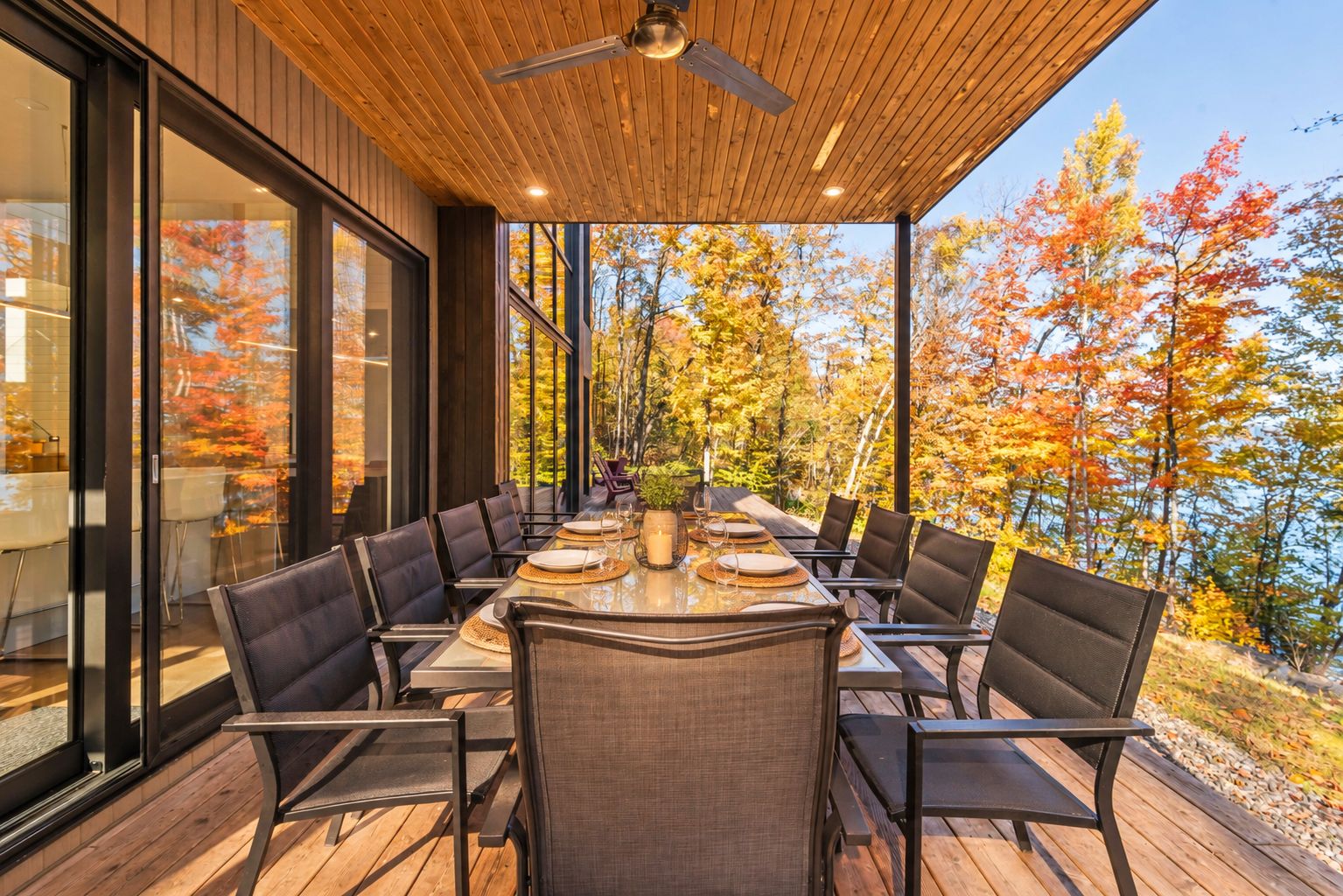An outdoor dining area with a wooden deck, large windows, and a view of autumn trees.