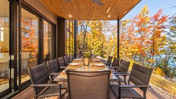 An outdoor dining area with a wooden deck, large windows, and a view of autumn trees.