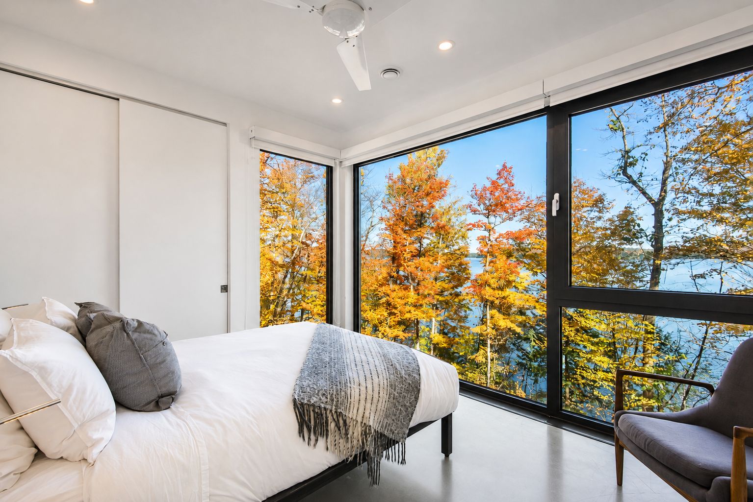 A bedroom with a large window, a bed with pillows, a bedside table, and a chair.