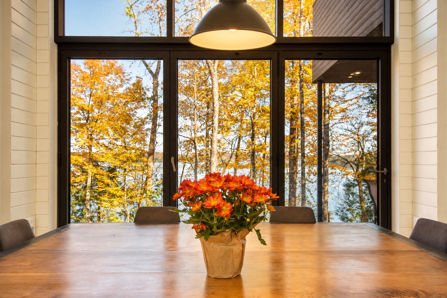 A dining area with a wooden table, chairs, a vase of flowers, and a view of autumn trees through large windows.