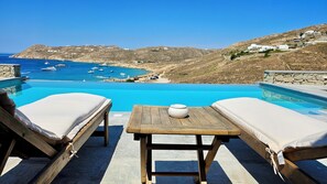 Uma área à beira da piscina com espreguiçadeiras e uma mesa, com vista para uma paisagem costeira com barcos e edifícios.