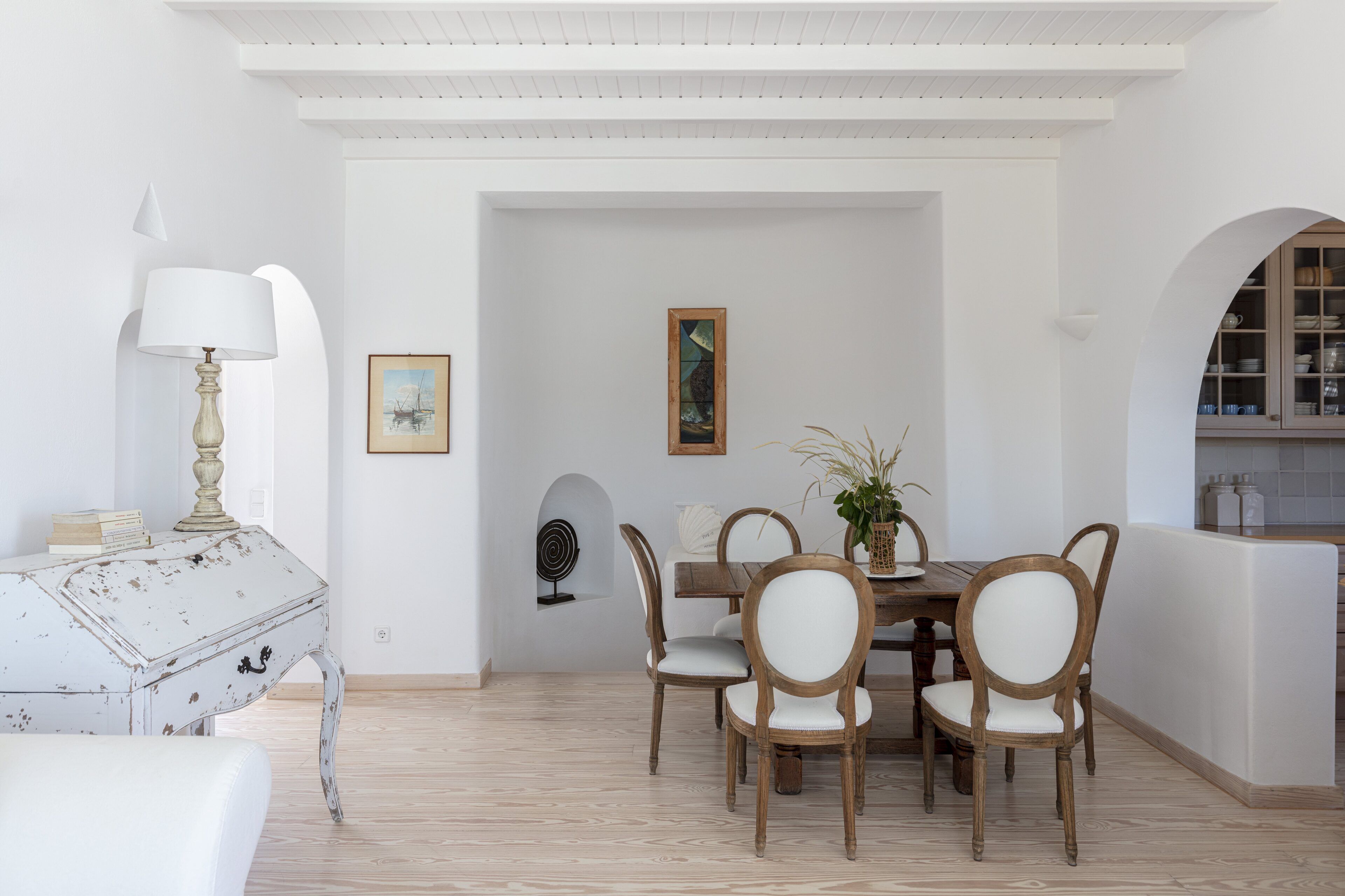 A dining area with a white table and wooden chairs, a white lamp, and a framed picture on the wall.