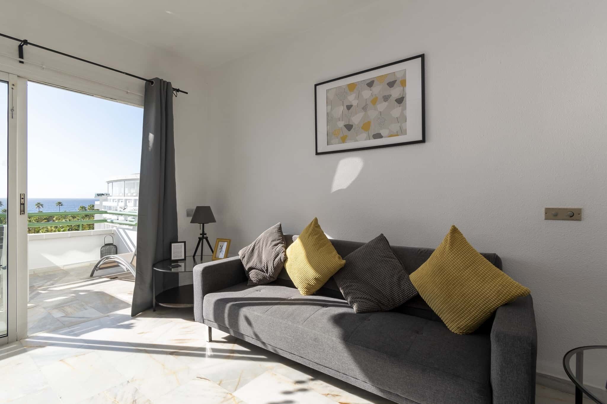 A modern living room with a grey sofa, a framed abstract painting, and a view of the sea through a sliding glass door.