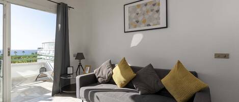A modern living room with a grey sofa, a framed abstract painting, and a view of the sea through a sliding glass door.