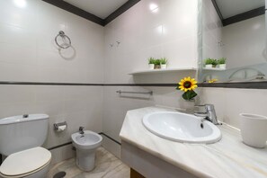 A modern bathroom with a toilet, bidet, sink, and mirror. There are two potted plants on a shelf above the sink.
