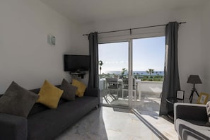 Relax in this stylish living room with a sofa, flat-screen TV, and sliding glass door leading to a furnished outdoor space with a scenic sea view.