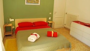 Un dormitori amb un llit, almoinades vermelles, un prestatge blanc, un radiador i un quadre a la paret.