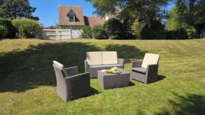 Un espace extérieur aménagé avec du gazon, des meubles en osier et une table, entouré d’arbres et d’un bâtiment dont le toit est recouvert de tuiles.