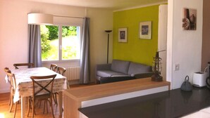 Un coin repas avec une table et des chaises, un canapé, une lampe sur pied, et un coin cuisine équipé d’un grille-pain et d’une bouilloire.