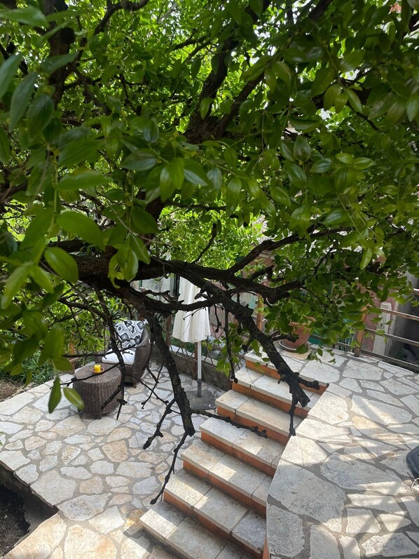 A stone patio with a tree providing shade, a wicker chair, and a small table. - Traditional Cottage and Garden Studio with Balcony, Terrace and Views to the Sea (Karousades)