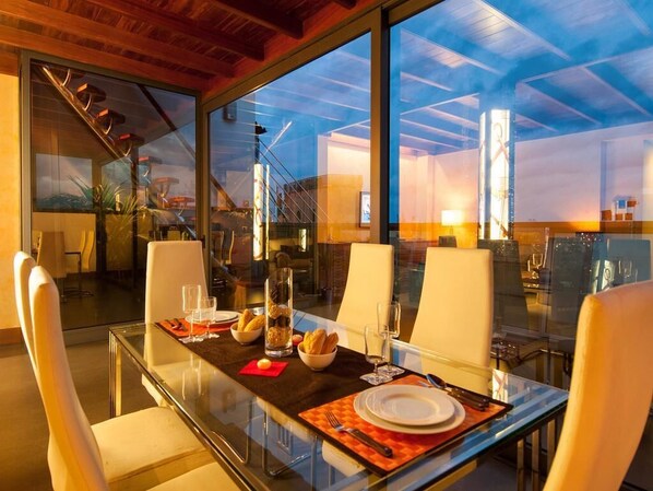 A modern dining area with a glass table, white chairs, and a view of the sky through large windows.