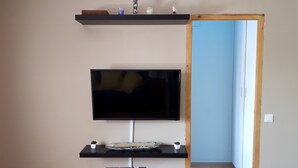Un salon moderne avec un téléviseur à écran plat fixé sur un meuble mural noir, une petite étagère au-dessus, et un cadre de porte en bois visible sur la droite.