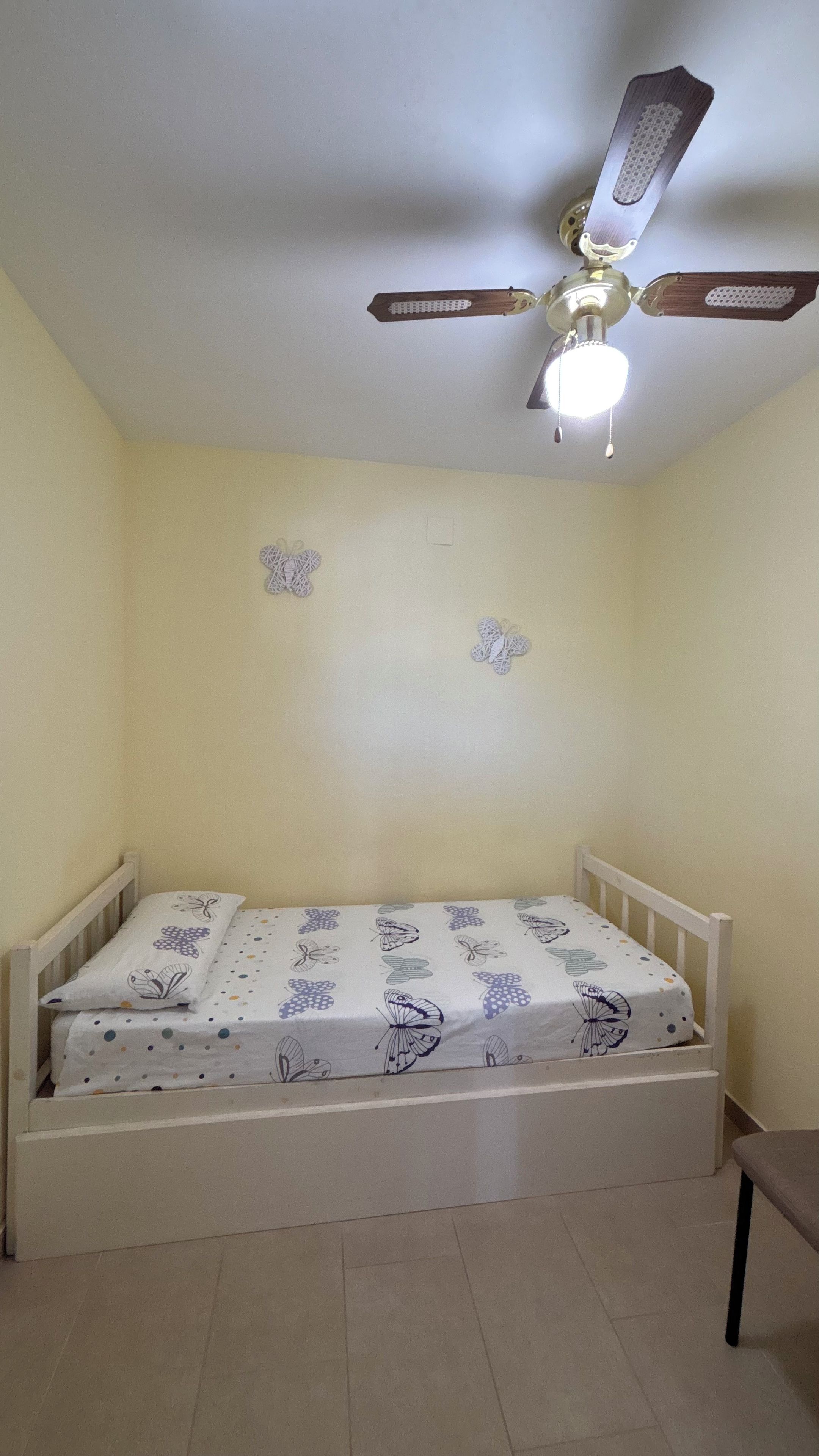A single bed with a white frame and a butterfly-patterned bedspread in a room with plain walls and a ceiling fan.