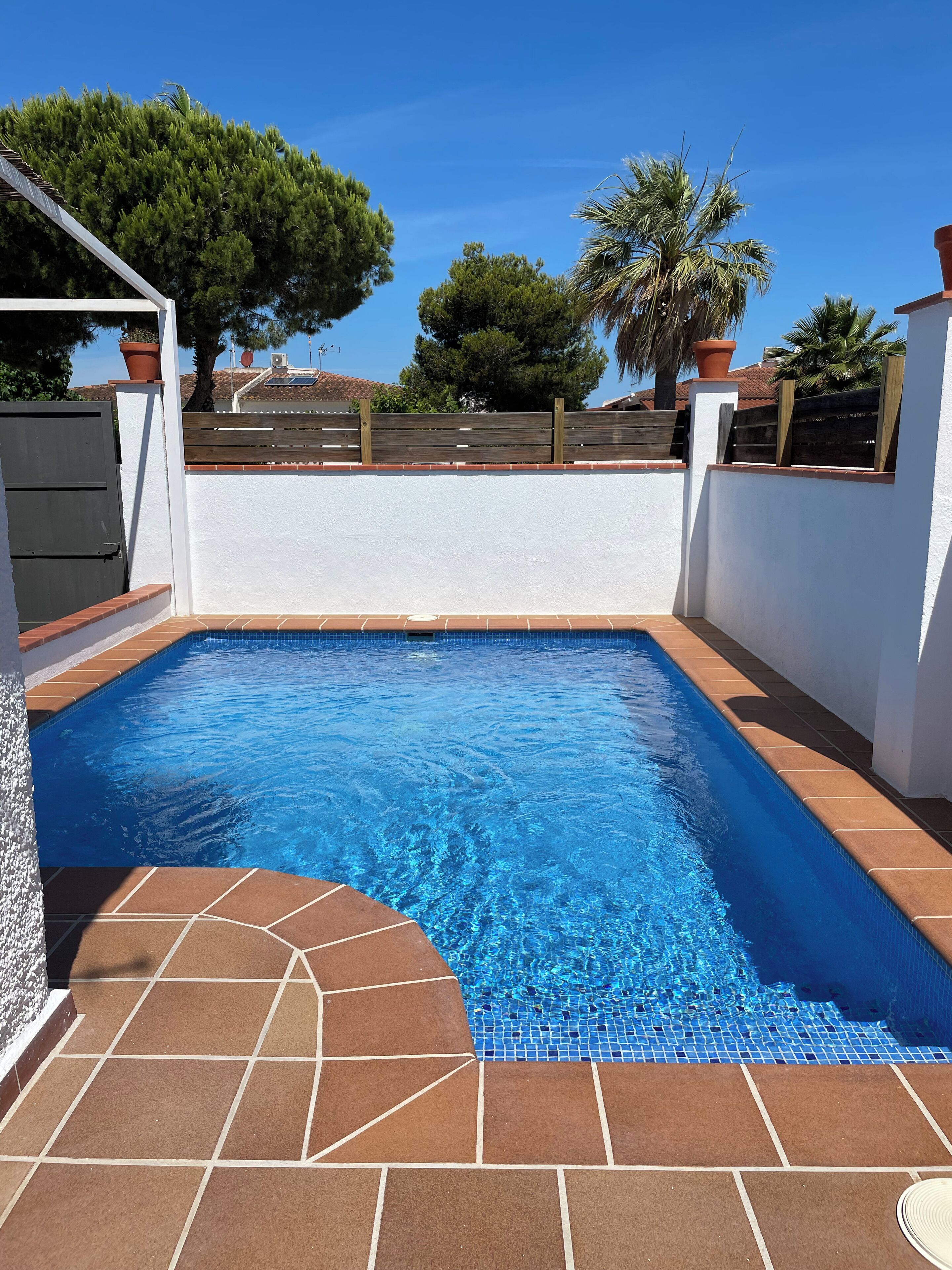 A rectangular swimming pool with clear blue water, surrounded by a white wall and a wooden fence.