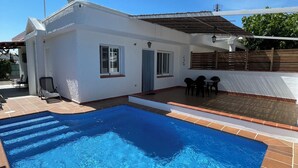 Une piscine résidentielle avec une terrasse carrelée, une maison blanche avec un toit en tuiles et un patio couvert.