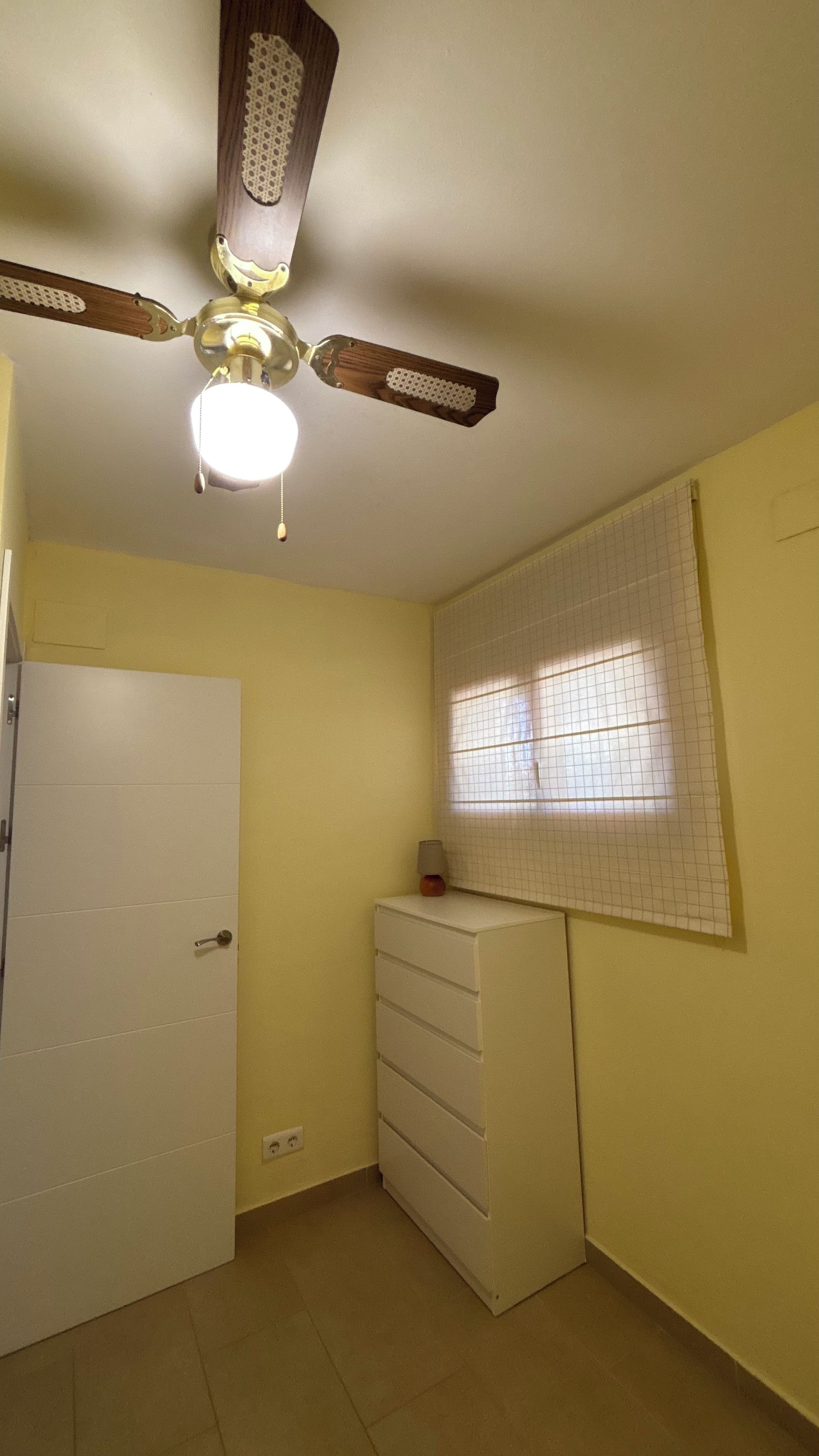 A room with a ceiling fan, a window with grid blinds, a white door, and a white dresser.