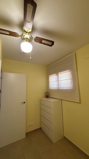 A room with a ceiling fan, a window with grid blinds, a white door, and a white dresser.