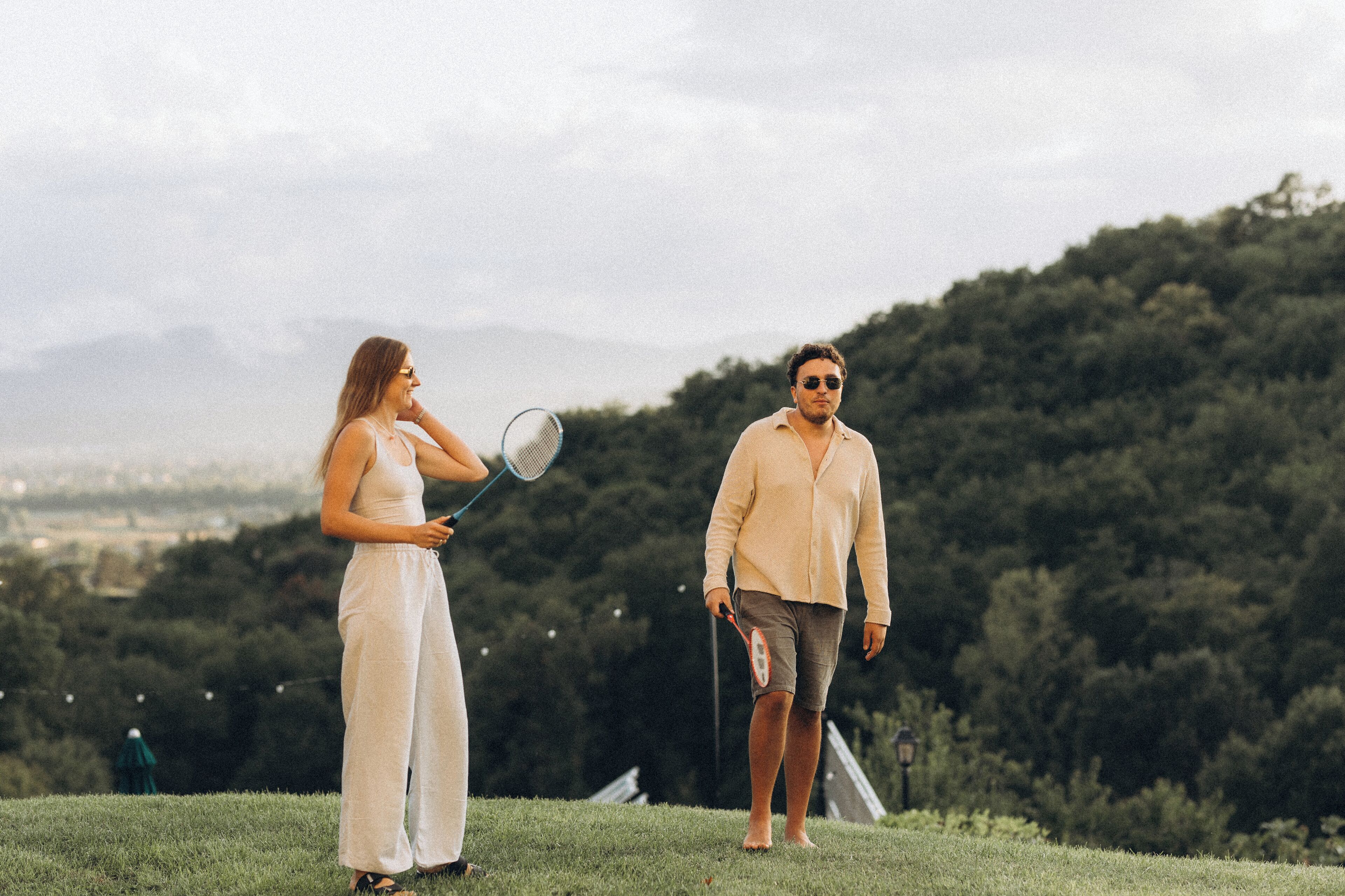 Two people standing on a grassy hill with a forested area and mountains in the background.